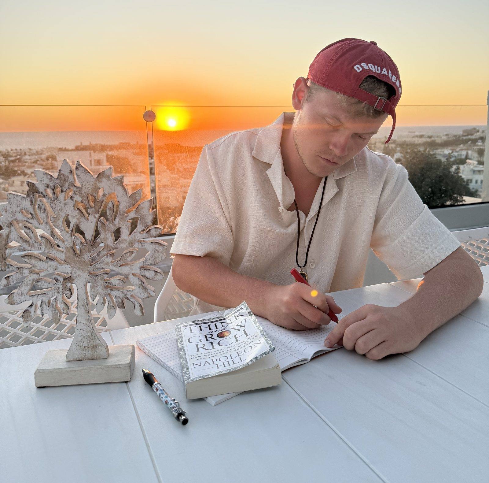 Niklas schreibt bei Sonnenuntergang in ein Notizbuch, neben ihm ein Buch und eine kleine Baumfigur.