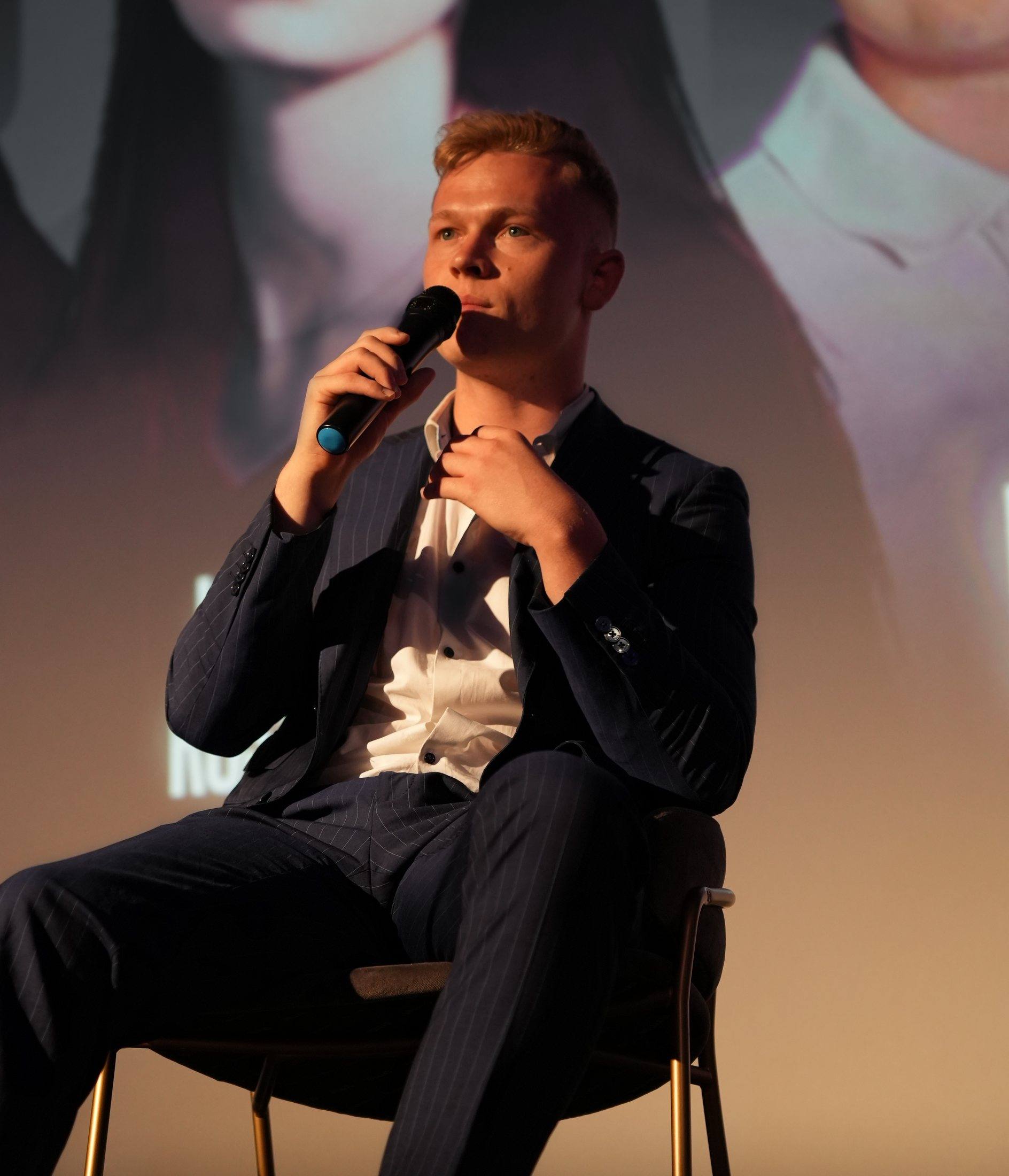 Niklas Benedikt Emtmann sitzt im Nadelstreifenanzug auf einer Bühne und spricht ins Mikrofon.