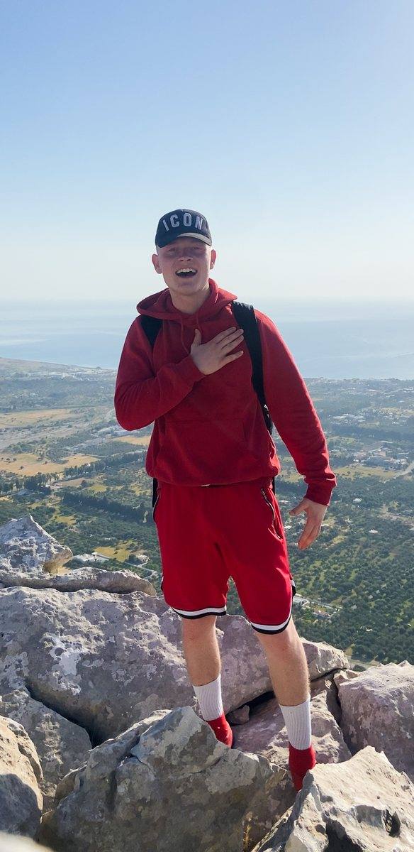Niklas steht im roten Kapuzenpullover auf einem Felsgipfel, hinter ihm Täler und Meer.