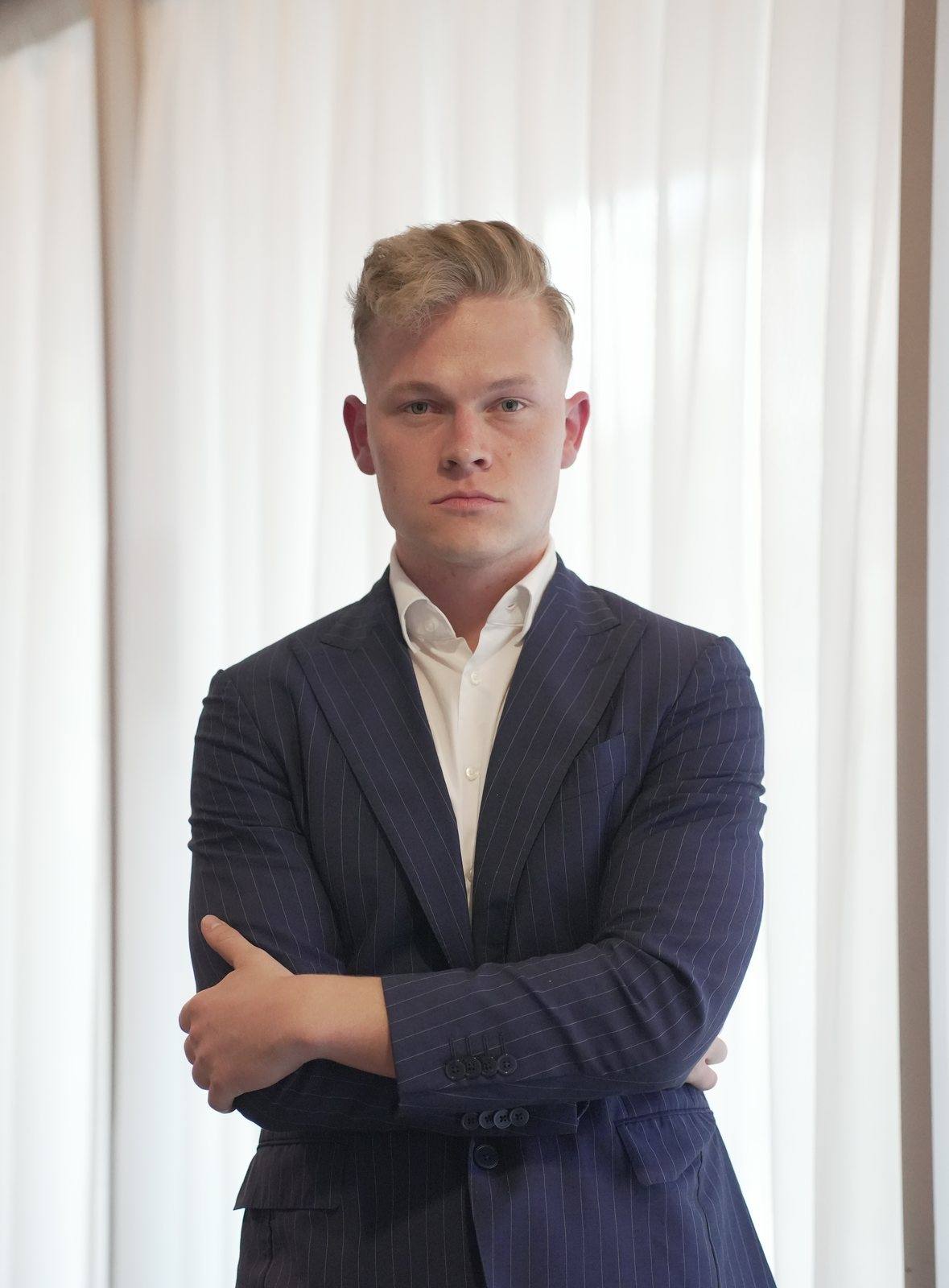 Niklas im dunkelblauen Nadelstreifenanzug vor hellen Vorhängen, die Arme verschränkt.