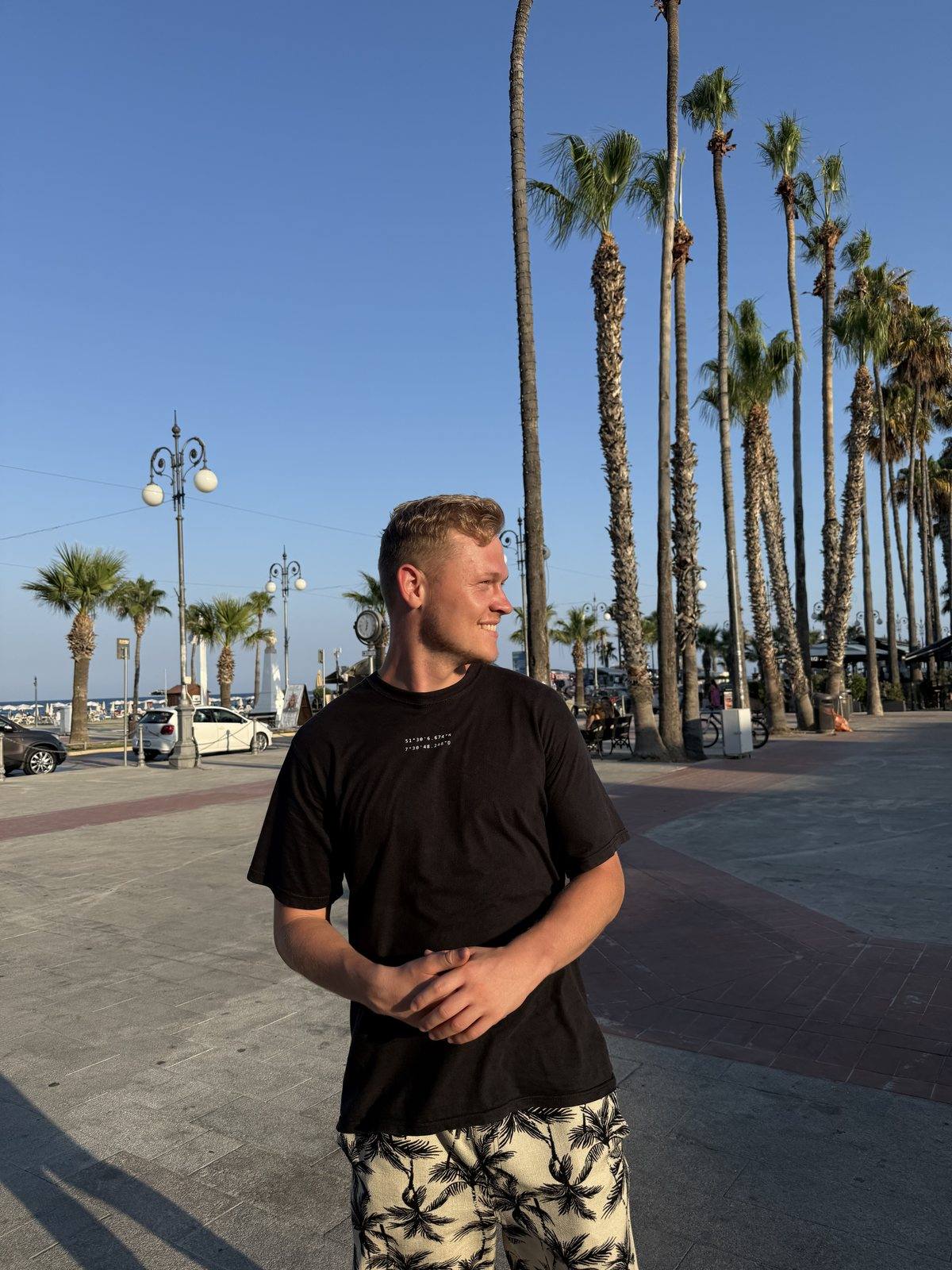 Niklas steht lächelnd an einer Promenade mit hohen Palmen unter blauem Himmel.